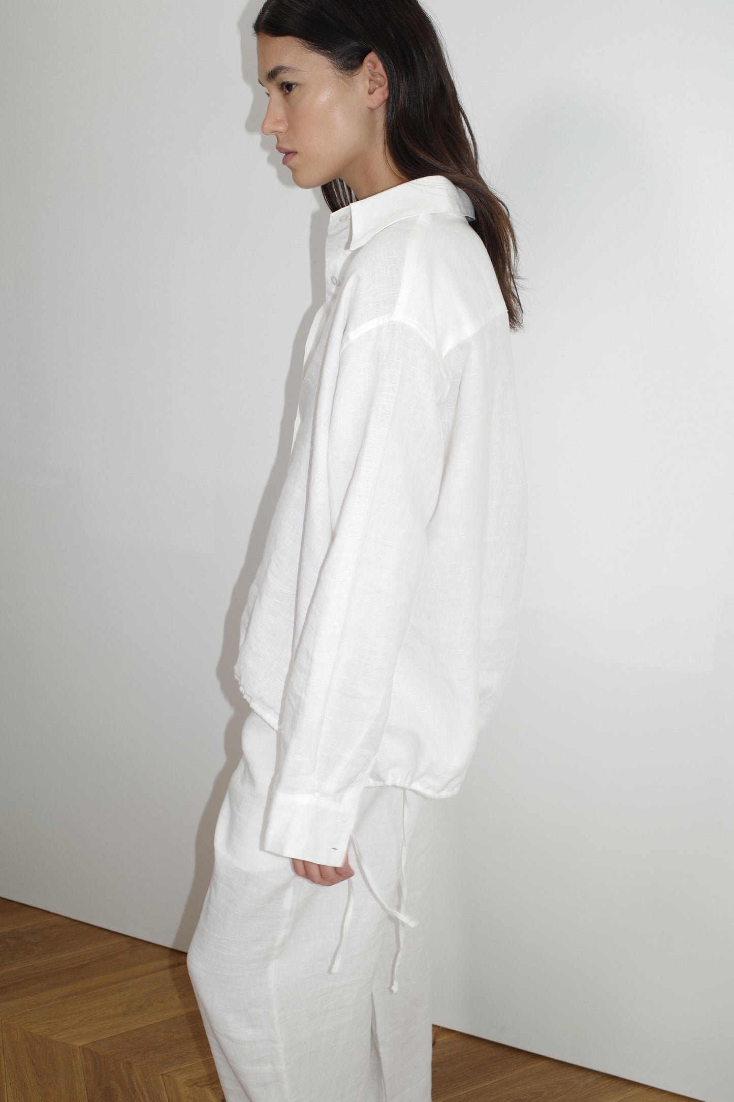 A woman with dark hair stands sideways against a white wall, wearing the Tie Split Set - White: a relaxed fit organic linen long-sleeve shirt and matching pants. She appears calm, standing on wooden flooring.