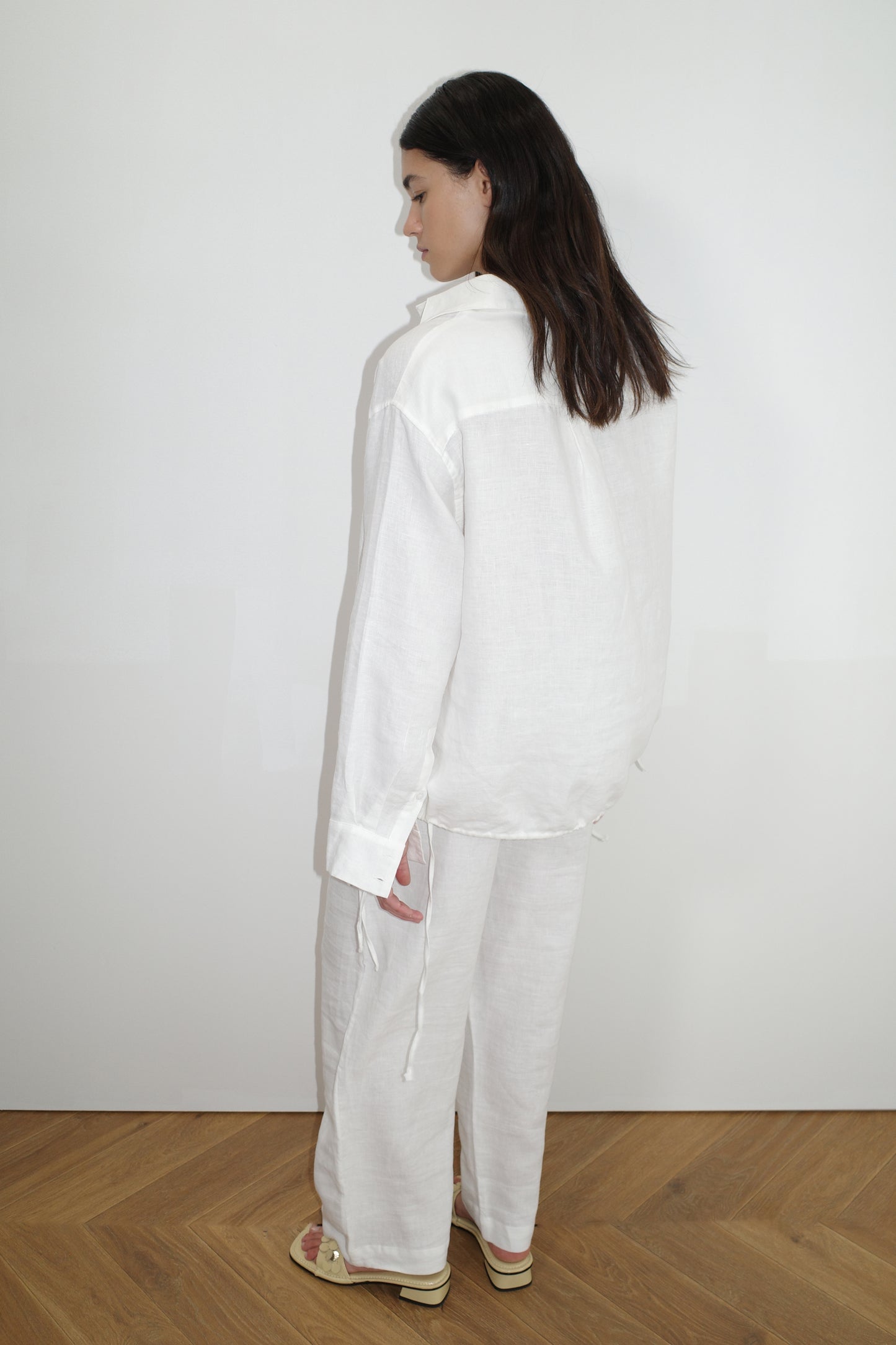 A woman with long dark hair stands sideways, gazing down. She wears the Tie Split Set - White: an airy, relaxed-fit organic linen shirt and matching wide-leg pants. Barefoot on a wooden floor, she poses against a plain white background.
