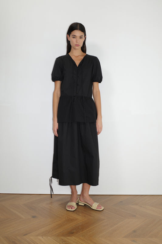 female model wears the draw hem dress by Deiji Studios in black
elevated everyday dress in mid length features a flattering v neckline with tie, button front detail with a drop waist, and soft elastic blouson sleeve
made from 100% organic cotton poplin