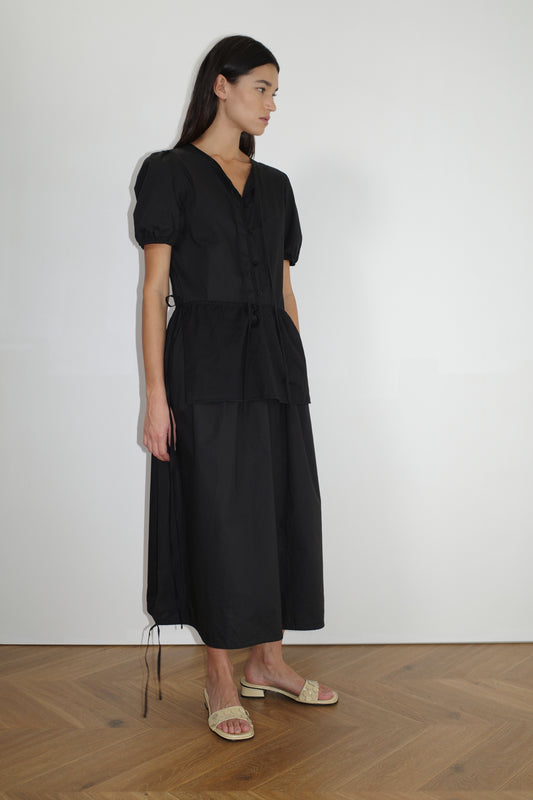 female model wears the draw hem dress by Deiji Studios in black
elevated everyday dress in mid length features a flattering v neckline with tie, button front detail with a drop waist, and soft elastic blouson sleeve
made from 100% organic cotton poplin