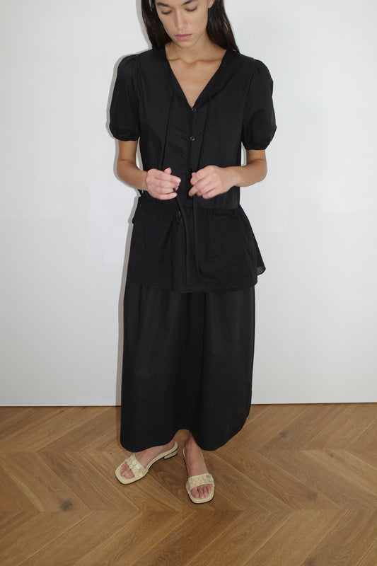 female model wears the draw hem dress by Deiji Studios in black
elevated everyday dress in mid length features a flattering v neckline with tie, button front detail with a drop waist, and soft elastic blouson sleeve
made from 100% organic cotton poplin
