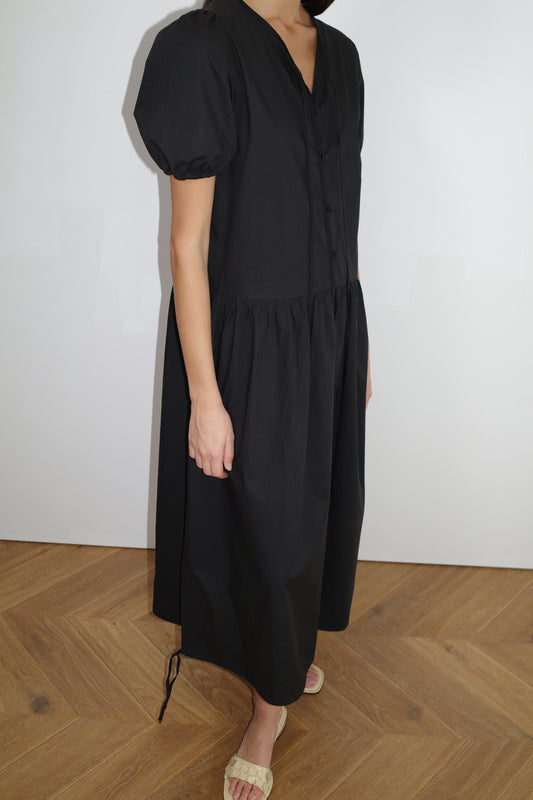 female model wears the draw hem dress by Deiji Studios in black
elevated everyday dress in mid length features a flattering v neckline with tie, button front detail with a drop waist, and soft elastic blouson sleeve
made from 100% organic cotton poplin