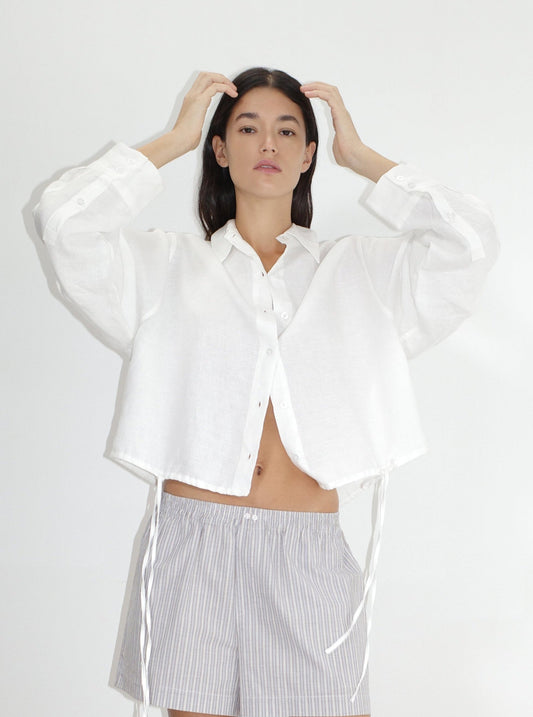 A woman wears the Hem Shirt - White by Deiji Studios, an oversized cropped French linen shirt with wide sleeves and ribbon ties, paired with grey striped shorts. She lifts her hands to her hair against a white background.