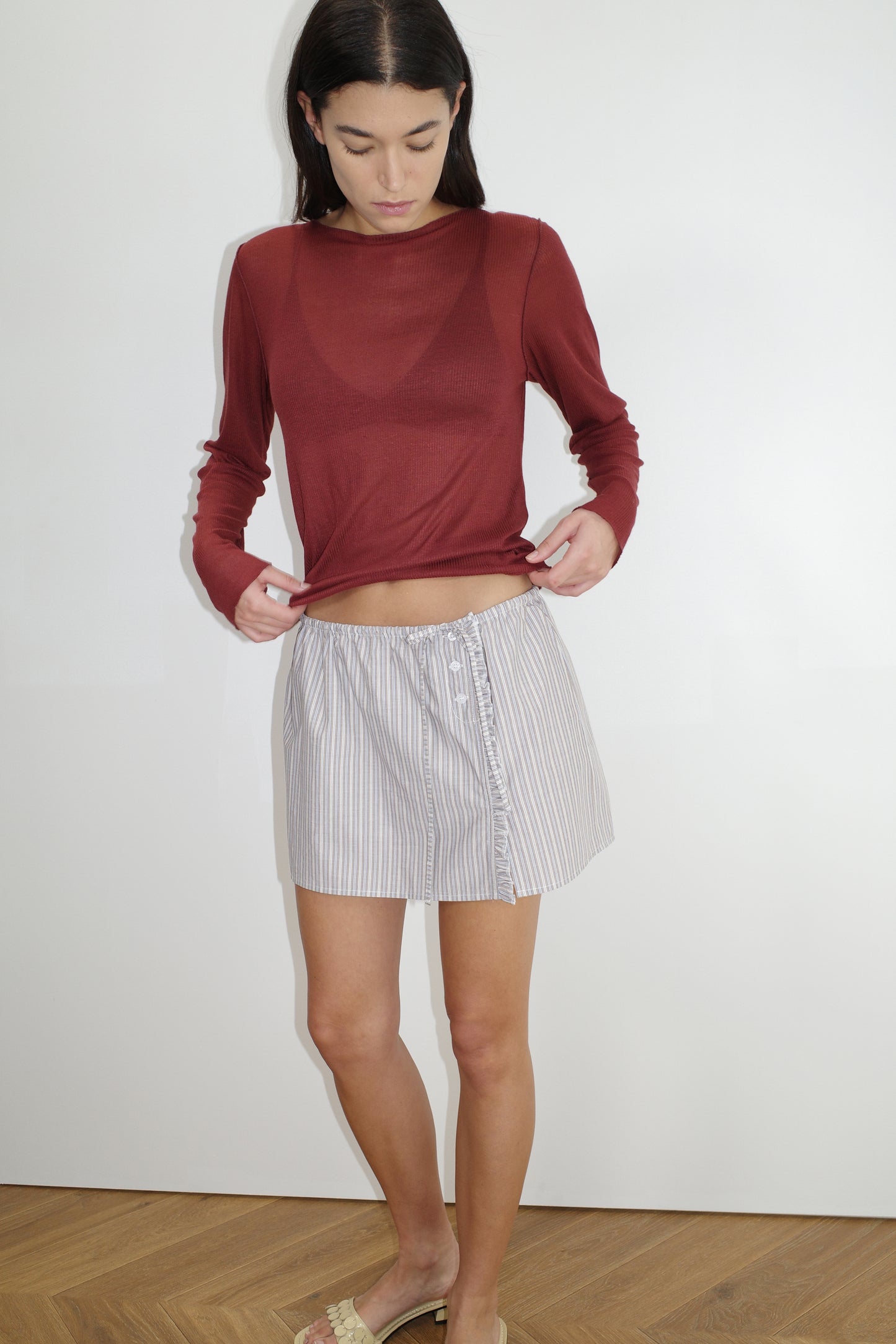 female model wears the ruffle mini skirt by Deiji Studios in seas stripe
simple mini skirt designed to sit at low waist features adjustable tie and ruffle edge on side with button detail
made from 100% GOTS certified organic cotton poplin
paired with a sheer knit long sleeve in crimson red