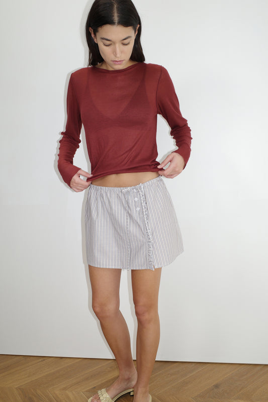 A woman stands indoors on a wooden floor, wearing a sheer maroon long-sleeve top and the Ruffle Mini Skirt. She looks down and slightly lifts the front of her top, with a plain white wall in the background.