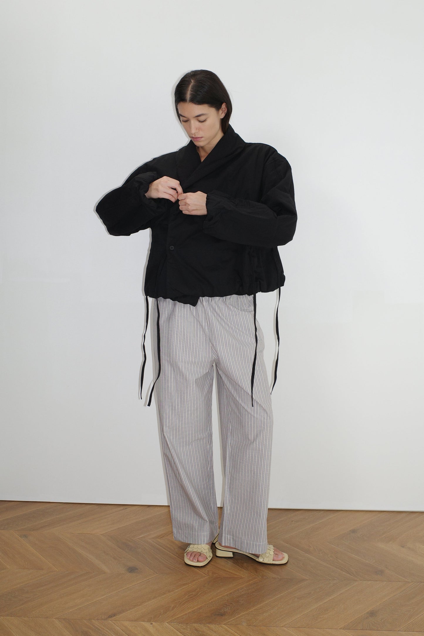 Indoors on a wooden floor, a person adjusts the long-sleeved Bubble Coat - Black by 0, worn over wide, light-striped organic cotton pants and sandals, standing against a plain white background.