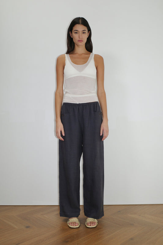 A woman with long dark hair stands indoors against a plain white wall, wearing the Linen Square Pocket Pant in dark gray, a sheer white tank top, and beige sandals, with her arms relaxed at her sides and a neutral expression.