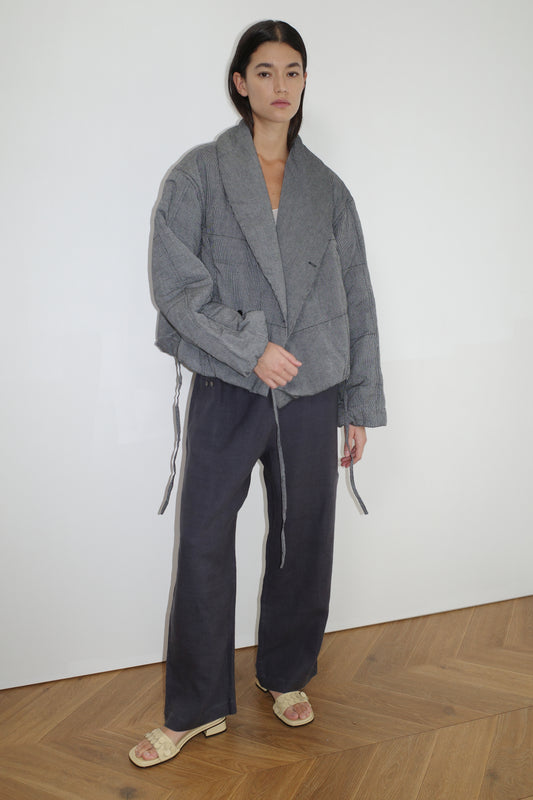 A woman with straight dark hair stands indoors against a white wall, wearing the 0 Exclusive Bubble Coat in Black White Stripe. She pairs it with wide-leg pants and cream slide sandals. Her outfit features soft organic cotton fabrics.
