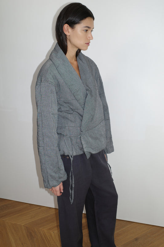 Someone with straight brown hair wears the Exclusive Bubble Coat - Black White Stripe by 0, paired with loose dark pants and an organic cotton top, standing indoors on a wooden floor against a plain white wall.