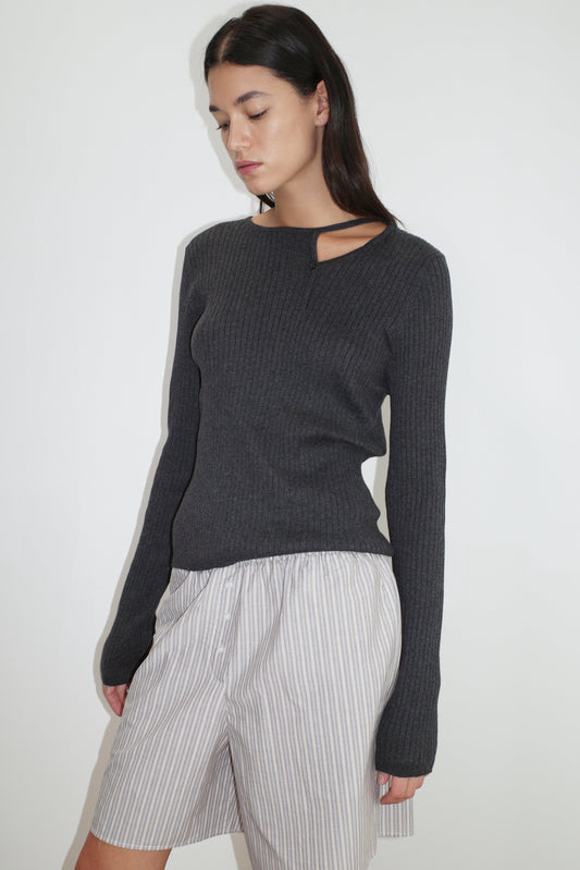 A woman wears the Cut Out Cardi - Coal by Deiji Studios, a dark gray ribbed knit sweater featuring a neckline cut-out. She pairs it with loose light-striped shorts and stands against a white background, gazing downward with a neutral expression.