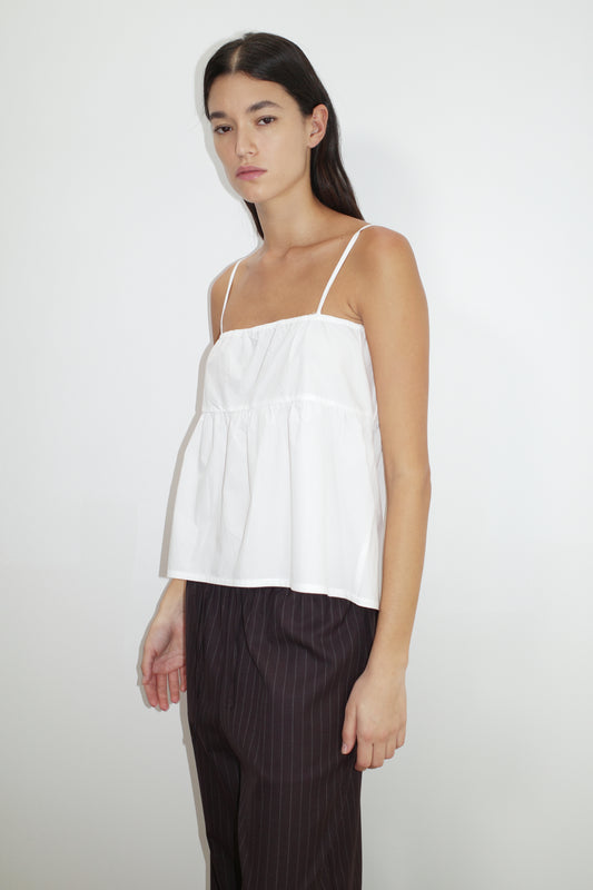 A person with long dark hair wears the Double Tie Top, a loose, sleeveless white top with thin straps, paired with dark pinstriped pants, standing against a plain white background.