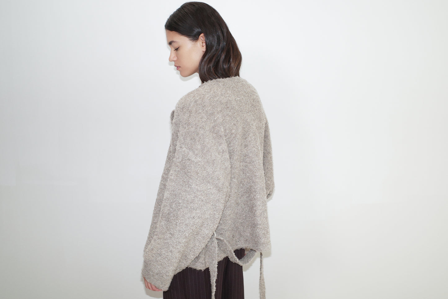 female model wears the pod knit by Deiji Studios in cloud grey
a relaxed and textured knit features a round shaped neckline with press stud front closure, removable self tie belt with long sleeves and high back neckline
made from a sustainable knit blend of 12% RWS certified wool, 30% recycled acrylic, 64% recycled polyester and 4% spandex
paired with a pinstripe wool pant