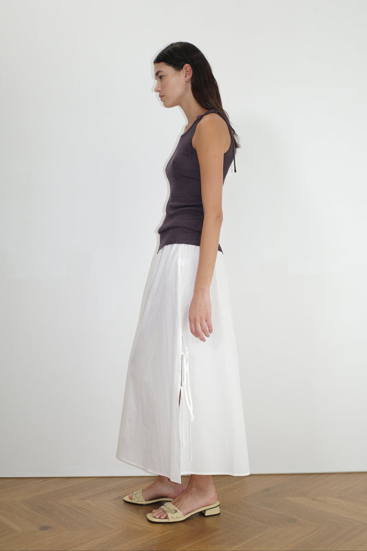 A woman stands in profile against a white wall, wearing a sleeveless dark purple top, the Split Tie Skirt in white with side ties, and beige slide sandals on a wooden floor.