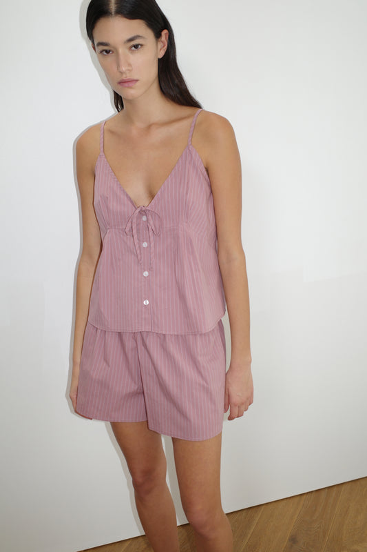 A woman with long dark hair and a neutral expression stands against a white wall wearing the Placket Short Set - Salt Stripe, featuring light purple pinstriped organic cotton with a sleeveless top, thin straps, buttons, and a small bow.