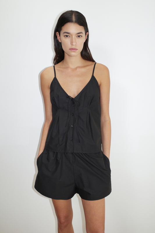 A woman stands against a white background, wearing the Placket Short Set—a black sleeveless romper with thin straps, front buttons, and pockets. She has a neutral expression and her hands in her pockets.