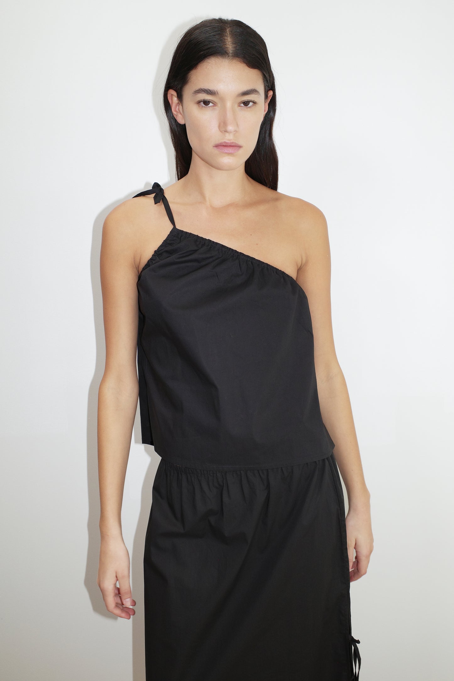 female model wears the ruched strap top by Deiji Studios in black
a lightweight one shoulder top featuring fine tie shoulder strap, fixed gathering on armholes and elasticated neckline for a relaxed fit
made from 100% organic cotton poplin
paired with a black skirt