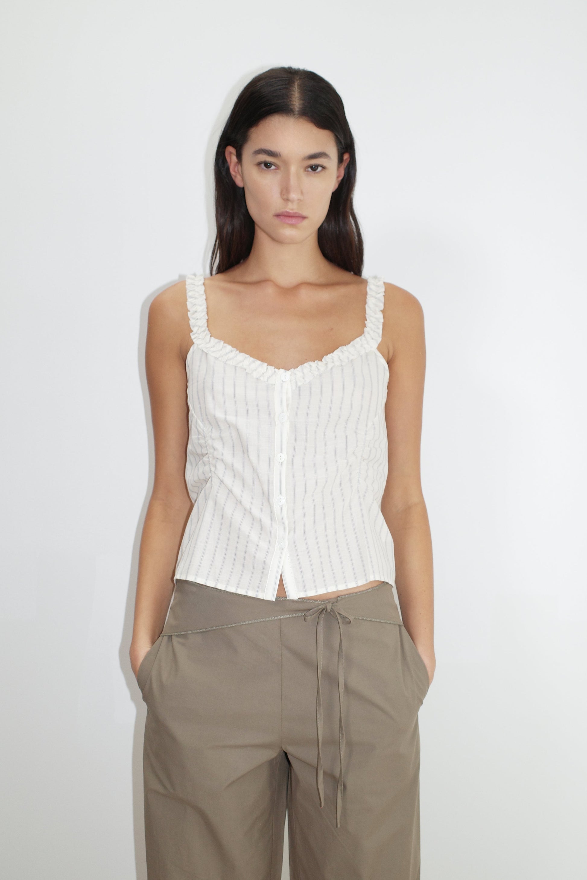 female model wears the ruffle button top by Deiji Studios in raquette stripe
a button up top featuring a butter base with mixed blue stripe, flattering v neckline, self-fabric ruffles along the straps and designed elasticated gatherings
made from 100% GOTS organic cotton
paired with a clay green pant