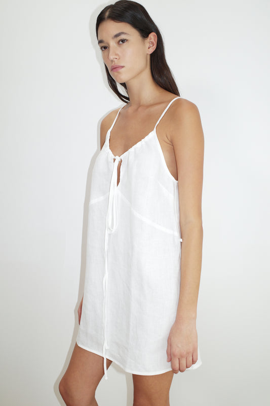 A woman with long dark hair and a neutral expression models the Tie Front Dress—a sleeveless, short white dress featuring thin straps and a chest tie—against a plain white background.