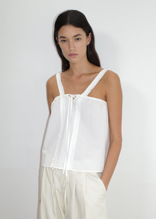 A woman with long dark hair wears the Ruffle Strap Top in white and cream-colored pants, standing against a plain white background with her hands in her pockets.