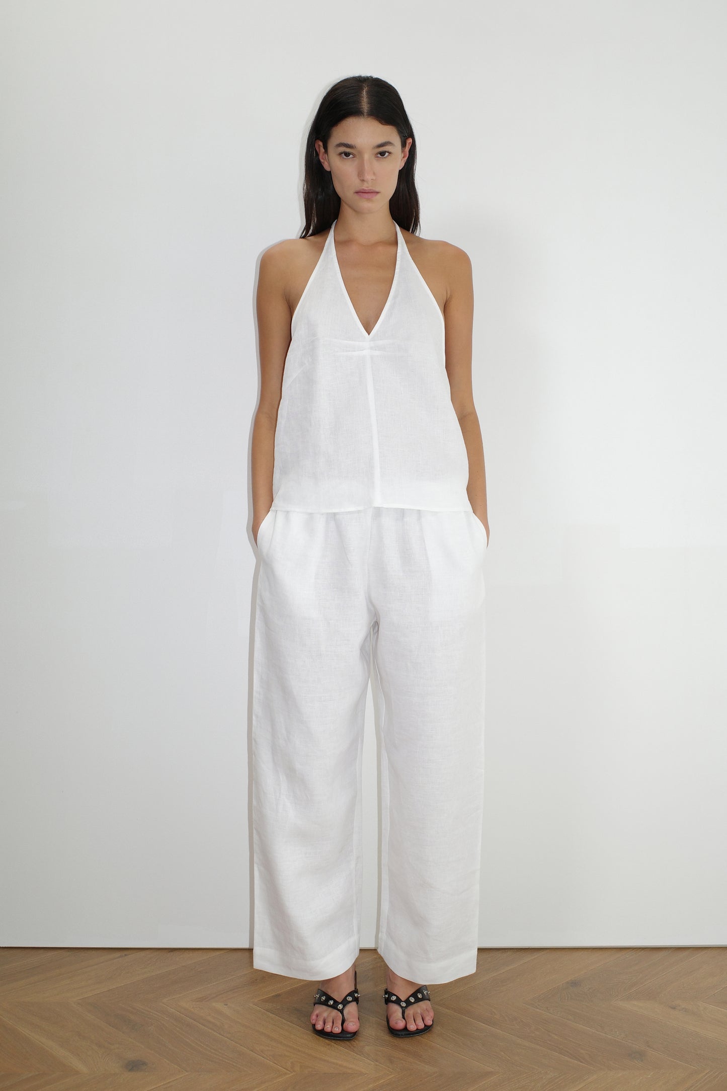 A woman stands against a plain white wall, wearing the Linen Pinched Halter Top - White with wide-legged white pants. She has straight dark hair, hands in her pockets, and completes the look with black sandals.