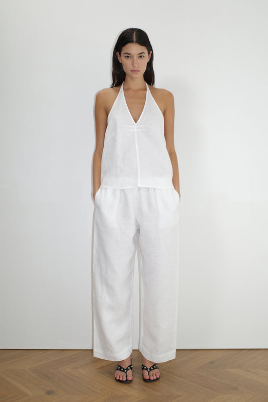 A woman with long dark hair stands indoors on a wooden floor wearing a sleeveless white halter top and the Linen Ease Trouser - White by Deiji Studios, paired with black sandals and her hands in the pockets.