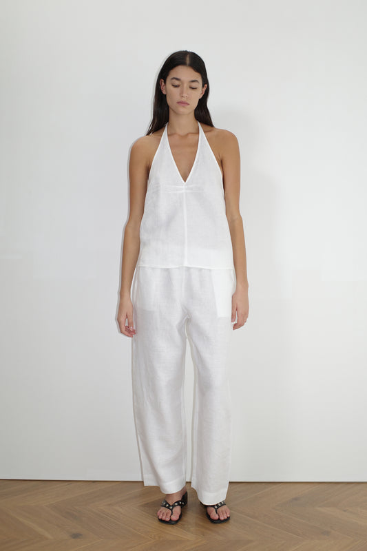 A woman with long dark hair stands indoors against a white wall, facing forward in a sleeveless white halter top, Linen Ease Trouser - White by Deiji Studios, and black sandals, wearing a neutral expression.