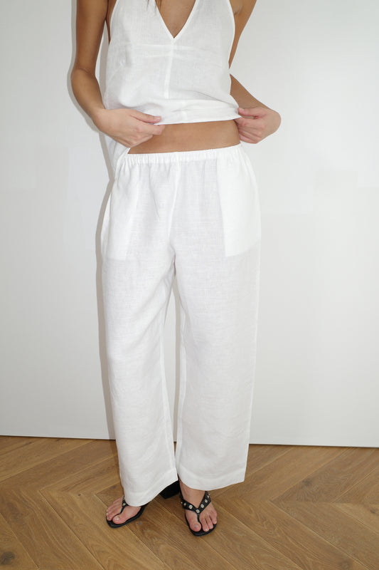 Wearing Deiji Studios' Linen Ease Trouser in White with a sleeveless white top, the person stands on a wooden floor holding up the top's hem. Face not visible, black flat sandals worn.