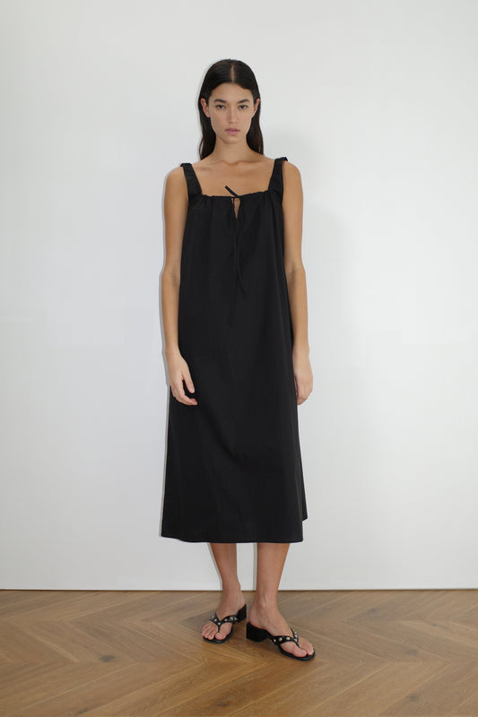 A woman with long dark hair stands indoors on a wooden floor, wearing The Paper Dress - Black by Deiji Studios—a loose, sleeveless maxi dress with a square neckline and bow tie detail—paired with black sandals, facing forward against a plain white wall.
