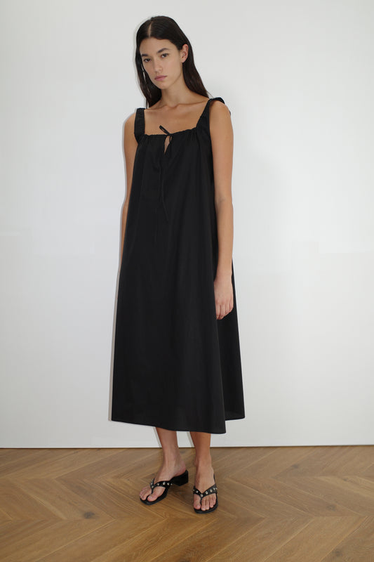 A woman with long dark hair and a neutral expression poses indoors on a wooden floor, wearing The Paper Dress - Black by Deiji Studios. The sleeveless, maxi-length dress features shoulder ties. She pairs it with black sandals against a plain white wall.