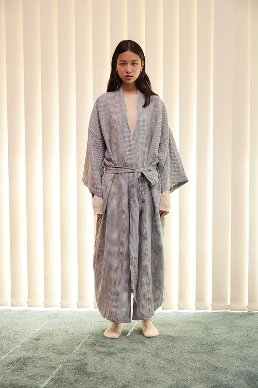 A person stands indoors on a green carpet, wearing The 02 Robe - Field Check by Deiji Studios—a full-length, gray striped robe made from stone washed French linen with a waist belt—paired with light socks, in front of cream vertical blinds.