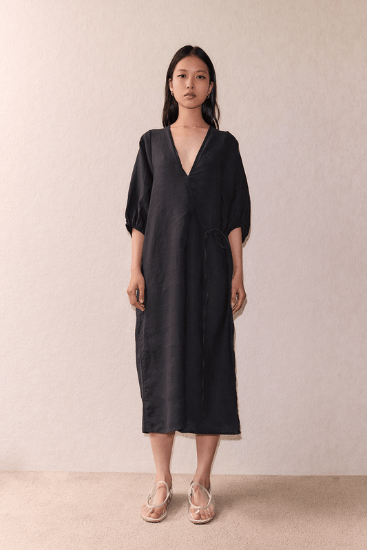 A woman with straight, shoulder-length hair and a neutral expression stands against a neutral background, wearing The Crease Bind Dress - Black by Deiji Studios, a loose V-neck wrap dress in dark linen with three-quarter sleeves and light flat sandals.