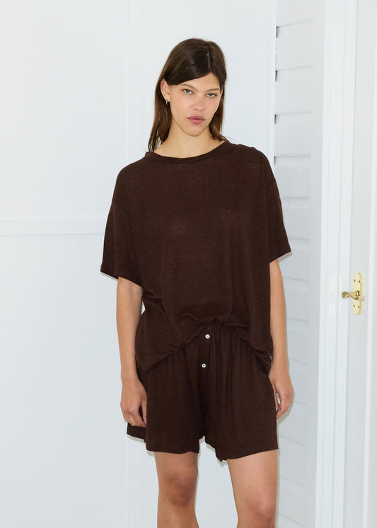 A woman stands indoors against a white wall, wearing the Soft T-Shirt in brown with matching shorts. She looks at the camera with a neutral expression.