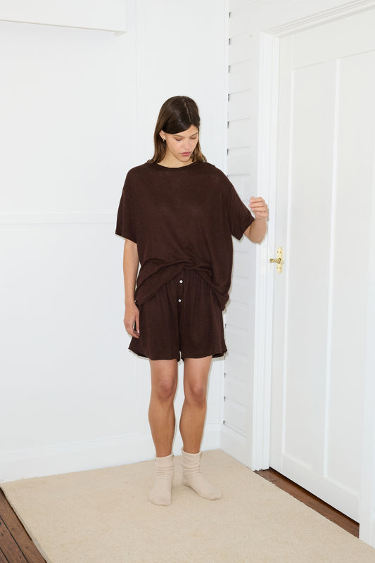 A woman stands indoors on a beige rug in a bright room, wearing the Soft Short with a matching loose brown top and white buttons, paired with beige socks. She looks down with one arm slightly raised against white walls and a door.