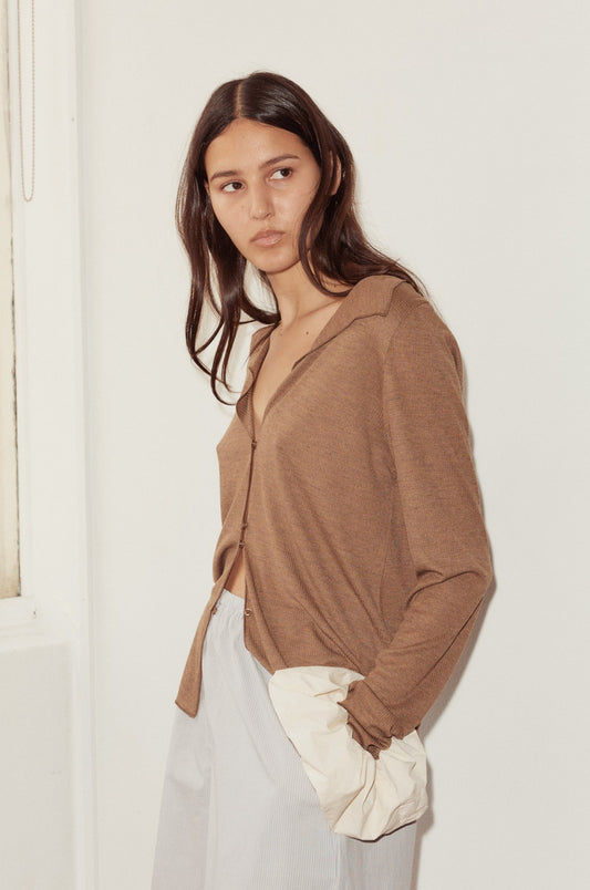 A woman with long brown hair stands indoors, wearing the Open Long Sleeve Top in light brown and light gray pants, one hand in her pocket, gazing slightly to the side with a neutral expression.