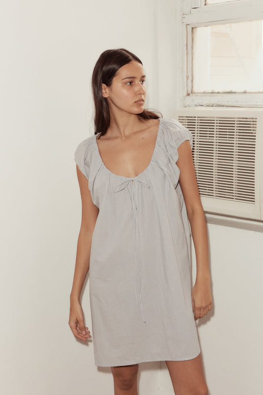 A woman with long brown hair stands indoors near a wall and window, wearing the Capped Sleeve Dress—a light gray, short-sleeved, loose-fitting style featuring a deep neckline and front tie detail.