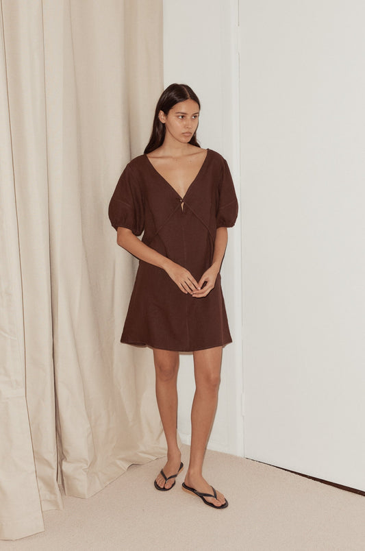 A woman wearing the Tie Seamed Short Dress—a short, loose-fitting brown dress with puffed sleeves—stands indoors on a beige carpet, near a white door and beige curtains, paired with black sandals.