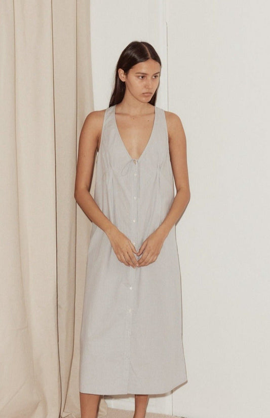 A woman with straight brown hair stands indoors in the Tuck Tie Dress - Dream Stripe, an organic cotton, sleeveless dress with a deep V-neckline. She faces slightly to the side, looking off-camera before neutral curtains and a white wall.
