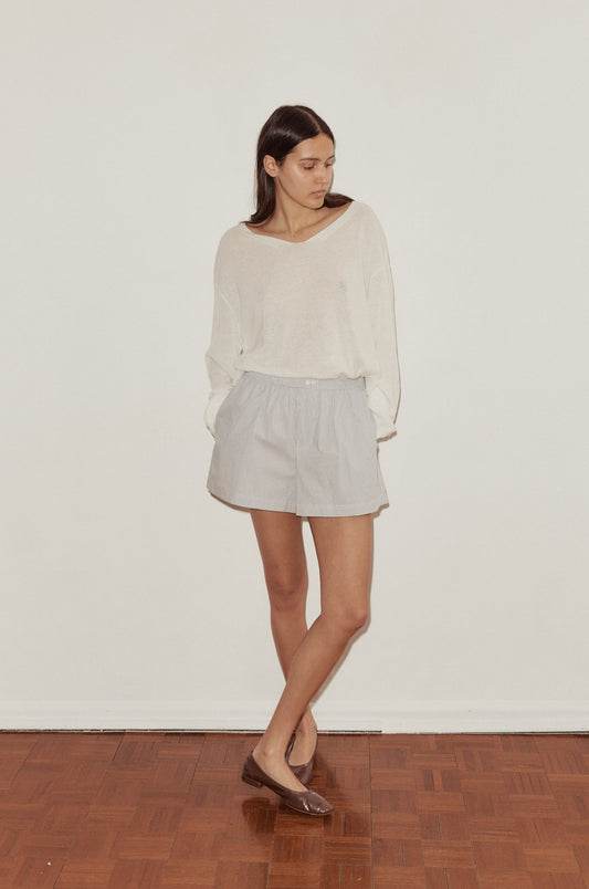 A woman stands indoors against a white wall, wearing the Organic Cotton Boxer in light gray, a loose long-sleeve top, and brown flats. She looks left with her hands in her pockets.