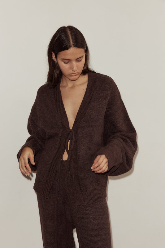 Wearing the Two Tie Front Cardigan, a person with long dark hair stands against a plain light background in matching knit pants, looking down with a relaxed expression.