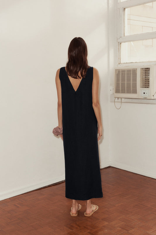 A woman stands on a parquet floor in a white room, seen from behind wearing the Keyhole Dress - Black, a sleeveless linen dress with a V-back neckline, paired with gold flat sandals.