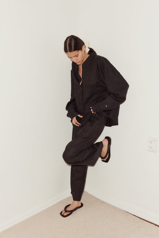 A woman poses in a corner wearing the Tack Set - Black—an oversized linen button-up and tapered trousers—paired with black flip-flops, a white scrunchie in her dark hair, and one leg raised.