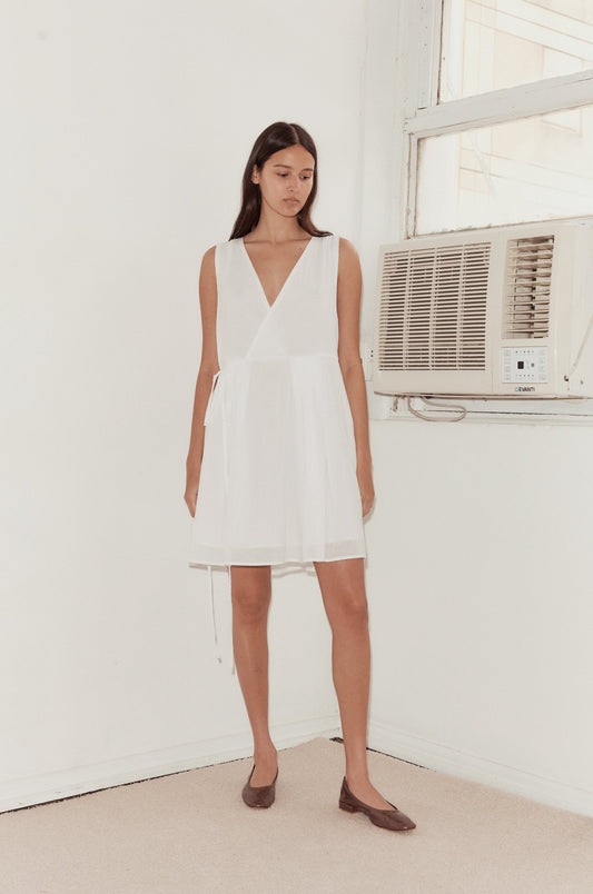 A woman in a sleeveless Wrap Tie Dress - White and brown flats stands indoors by a window with a wall-mounted AC, against a plain white wall and beige carpeted floor.