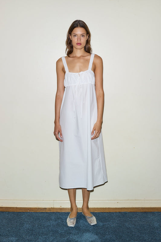 A woman with loose hair stands barefoot on a blue carpet against a light wall, wearing the Ruched Tie Dress—a sleeveless, calf-length white dress featuring thick straps and a gathered bodice.
