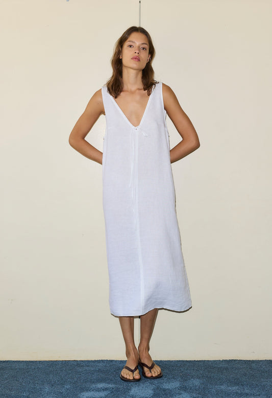 A woman stands against a beige wall, wearing the Tie Slip Dress—a loose, sleeveless white dress with a deep V-neck—paired with black flip-flops. Her arms are behind her back as she stands on a blue carpet.