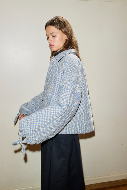 A person with long brown hair wears a light gray Collared Quilt Coat with puffy sleeves and cuff ties, paired with a long black skirt, standing against a plain light-colored wall.