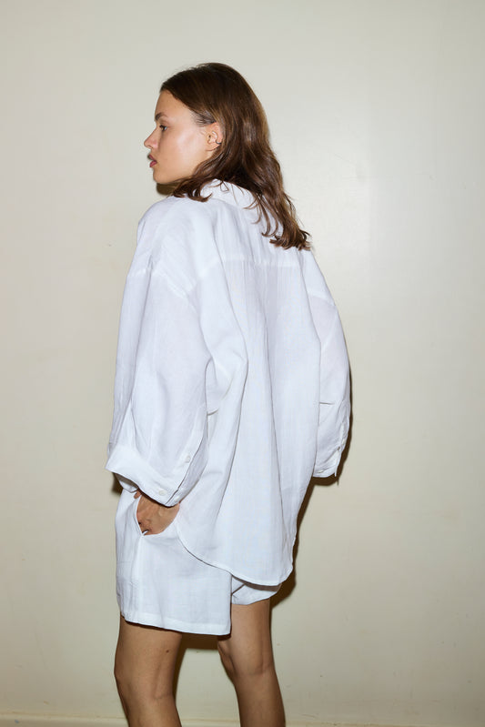A woman with medium-length brown hair stands sideways against a plain light wall, hands in her pockets, wearing the 03 set - white by Deiji Studios: an oversized linen shirt and matching white boxer shorts.