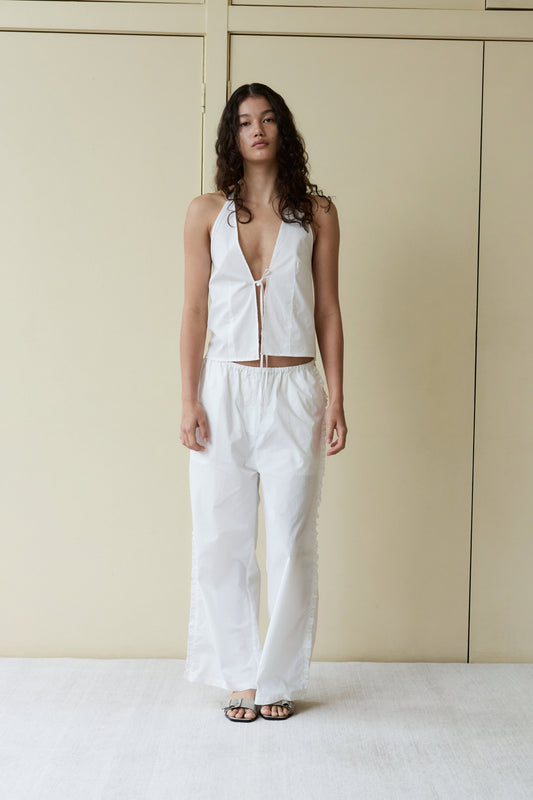 A person with long curly hair wears a sleeveless white halter top with a deep V-neck and loose Ruffle Pant, standing against a beige background and wearing black sandals.