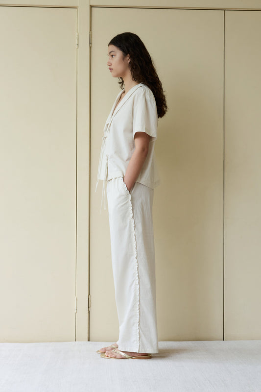 Woman in profile wearing Ruffle Pant—cream linen, wide-leg with side ruffle detail—paired with a matching wrap top and gold sandals, set against a neutral beige paneled background.