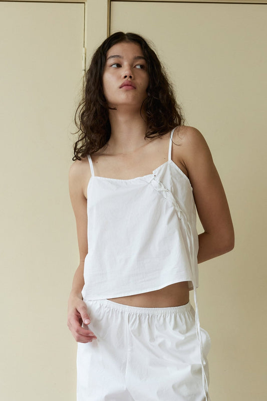 A young woman with wavy brown hair stands against a beige door, looking slightly to her left with a relaxed expression, wearing the Eyelet Top and matching white shorts.