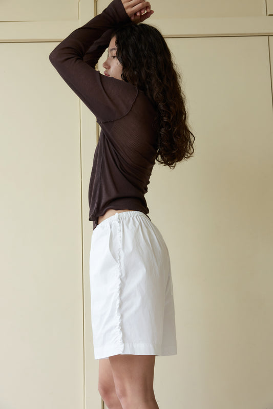 A person with long, curly hair stands in profile, wearing a sheer brown long-sleeve top and our white Ruffle Short, arms raised above their head against a beige background.